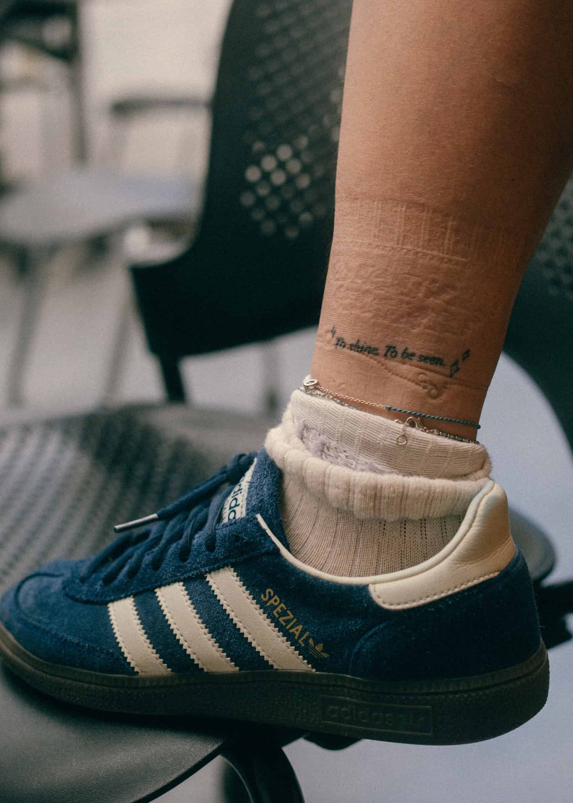 A close-up of a person's lower leg showing a tattoo with text, wearing white socks and blue Adidas Spezial sneakers, resting on a perforated black chair.