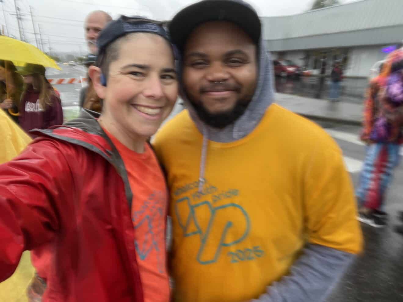 Two people wearing colorful shirts and jackets smile for a selfie at an outdoor event on a rainy day. Other participants and umbrellas are visible in the background.
