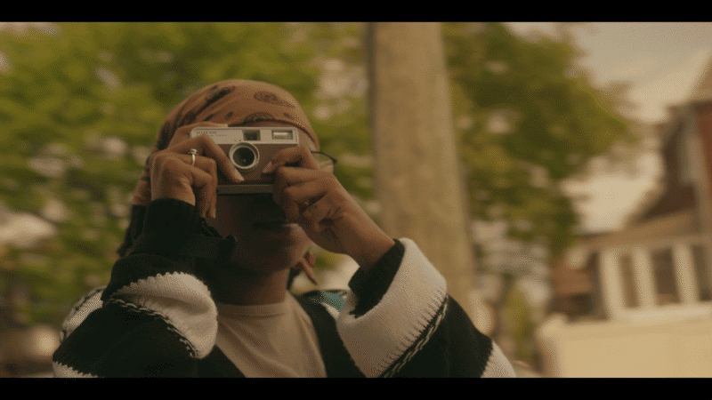 Person wearing a headscarf and striped sweater holds a camera up to their face, taking a photo outdoors with blurred trees and houses in the background.