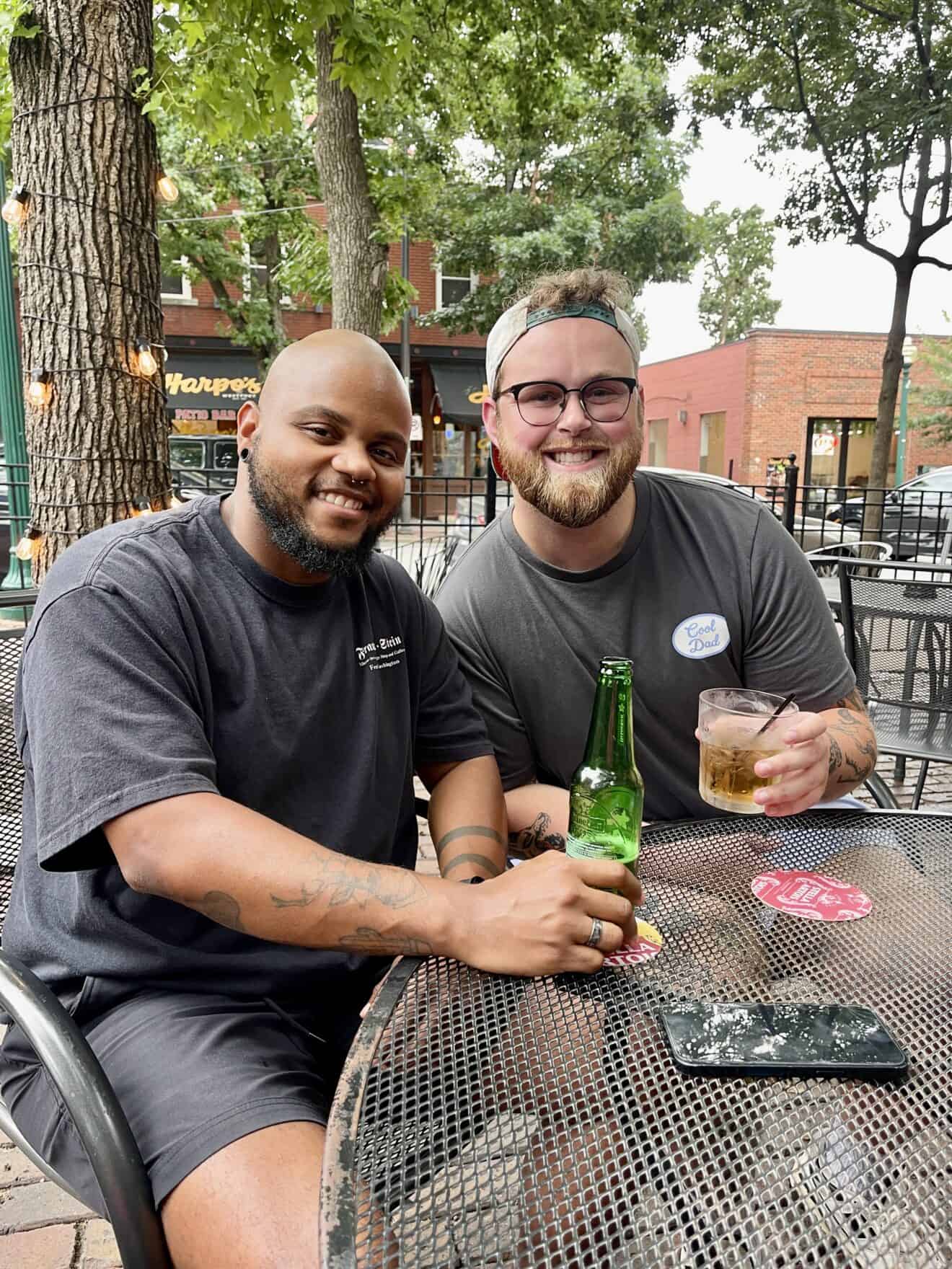 Two men sit at an outdoor metal table, smiling at the camera while holding drinks. Trees, string lights, and brick buildings are visible in the background.