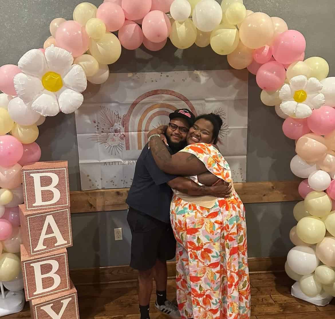Two people hug and smile under a pastel balloon arch with flower shapes at a baby shower, next to large blocks spelling "BABY.