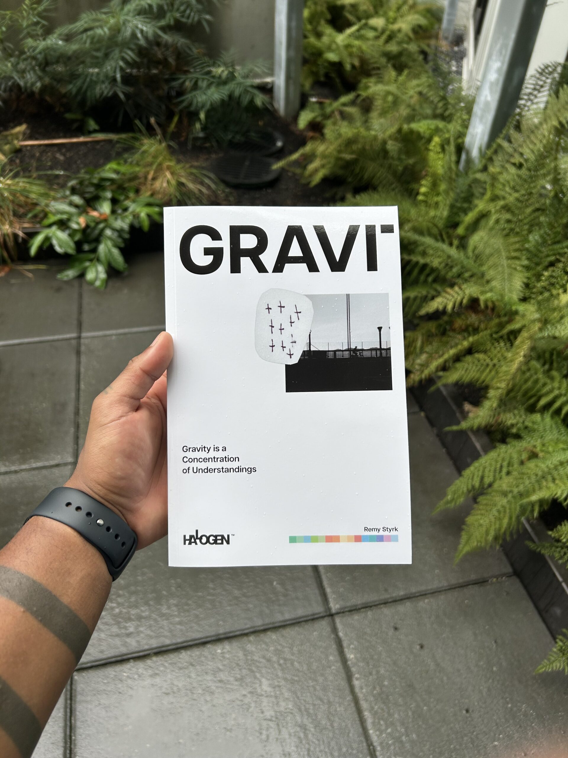 A person holding a book titled “GRAVI,” with a subtitle "Gravity is a Concentration of Understandings," standing on a patio surrounded by plants and ferns.