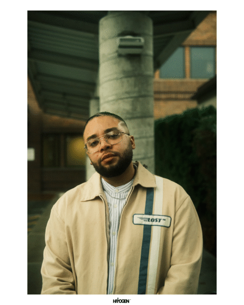 A man with glasses and a beard stands outdoors in front of a building, wearing a beige jacket with a "LOST" patch and a striped shirt.
