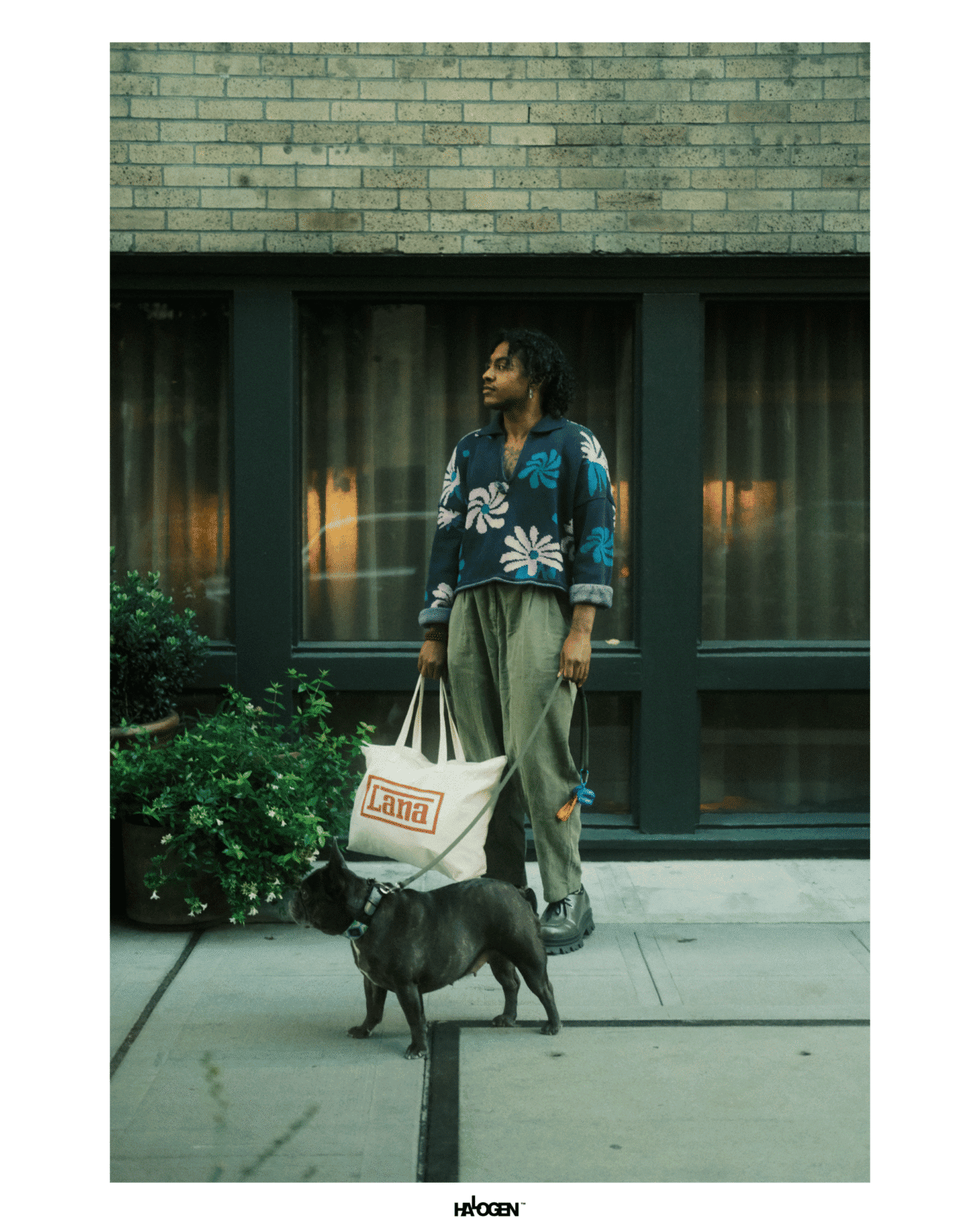 Person standing on a sidewalk holding a large tote bag and leash, with a small black dog beside them, in front of a brick building with large windows.