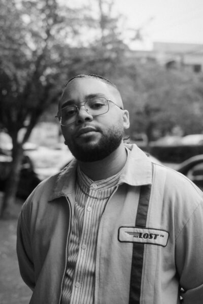 A man with glasses and a beard stands outdoors wearing a striped shirt and a jacket with a "LOST" patch; trees and cars are visible in the blurred background.