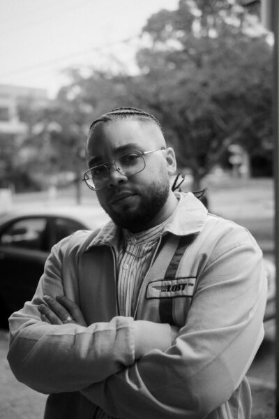 A person with glasses, a beard, and braided hair stands outdoors with arms crossed, wearing a light jacket over a striped shirt.
