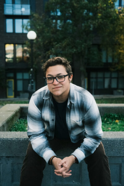 A young person with short brown hair and glasses sits on a concrete ledge outdoors, wearing a plaid shirt and dark pants, with trees and buildings in the background.
