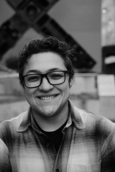 A person with glasses and short, wavy hair smiles at the camera while wearing a plaid shirt. The background features an abstract outdoor sculpture.