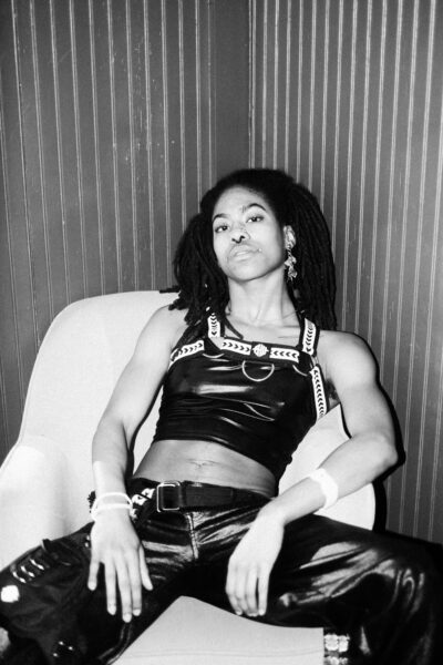 A person with long dreadlocks sits relaxed in a chair, wearing a sleeveless leather top, leather pants, and accessories, against a paneled wall.