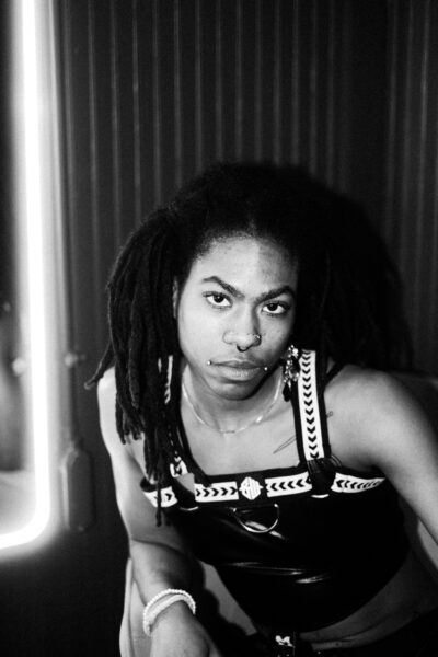 A person with long dreadlocks, a piercing, and facial glitter wears a strappy top and sits, looking directly at the camera in a black-and-white setting.