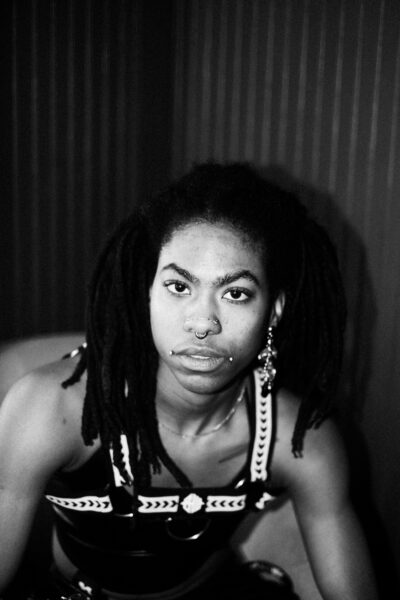 A person with long dreadlocks, facial piercings, and wearing a strappy outfit looks directly at the camera in a black and white photo.
