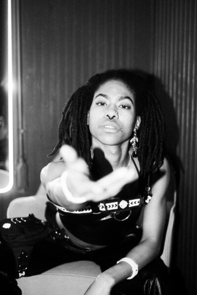 Person with dreadlocks and facial piercings sits on a chair, reaching one hand toward the camera, wearing dark clothing and accessories, in a dimly lit setting.