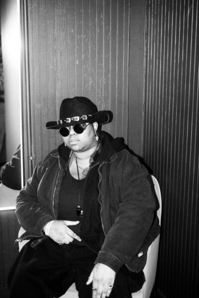 Person wearing a wide-brimmed hat, sunglasses, and layered clothing sits on a chair against a paneled wall, facing the camera with a neutral expression.