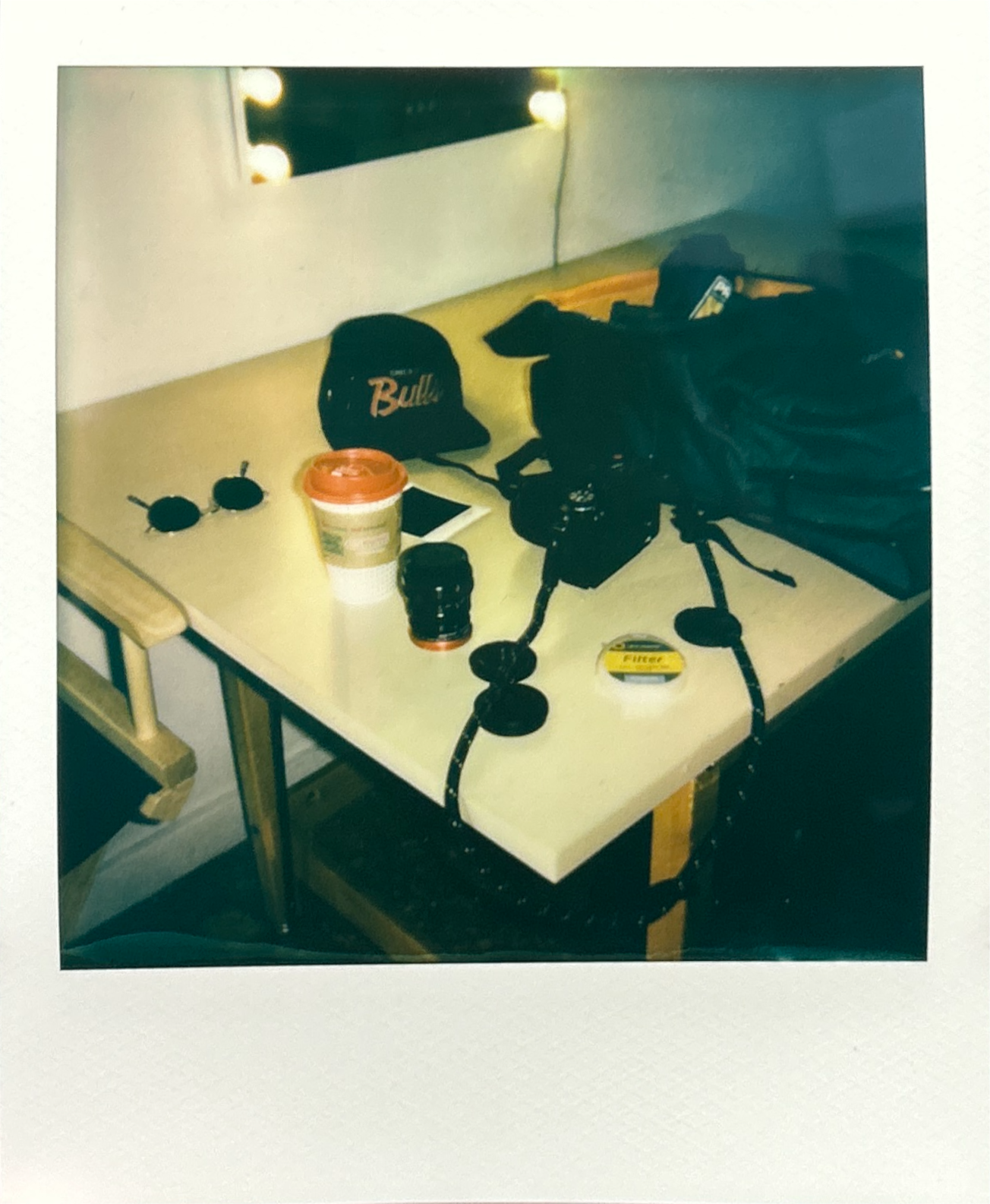 A table with sunglasses, a coffee cup, a camera, camera lens caps, a film canister, a cap, a phone, and a backpack near a mirror with lights.