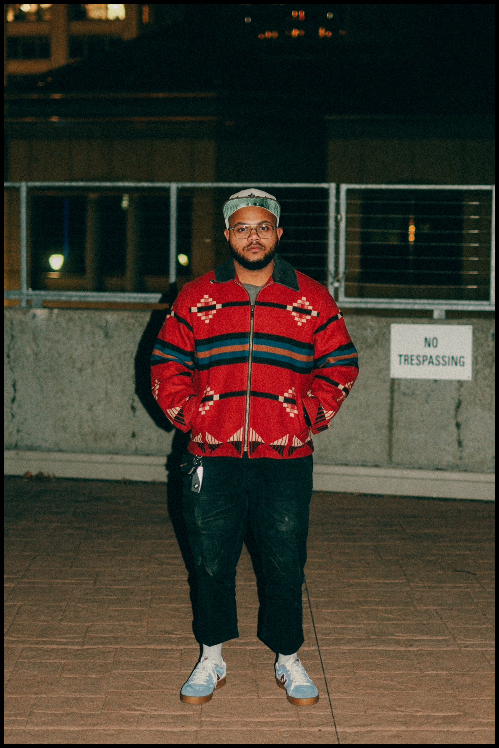 A man stands outdoors at night wearing a red patterned jacket, black pants, white sneakers, and a cap. A "No Trespassing" sign is visible on the wall behind him.