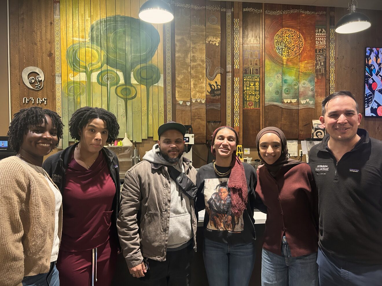 Six people stand together indoors, smiling at the camera in front of a wooden wall decorated with colorful, abstract art panels.