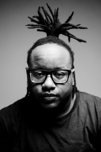 Black and white portrait of a person with glasses, a nose ring, and dreadlocks tied up, looking directly at the camera with a neutral expression.