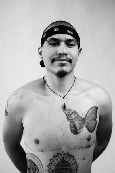 A shirtless person with a bandana, necklace, and various tattoos, including a butterfly on the chest, stands against a plain background.