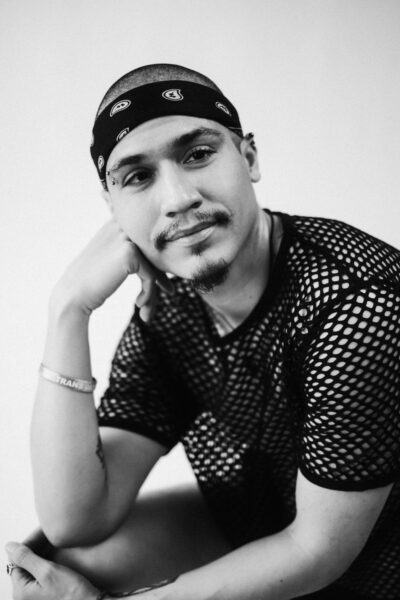 A person wearing a mesh shirt and bandana sits with one hand on their chin, looking slightly to the side. The photo is black and white.