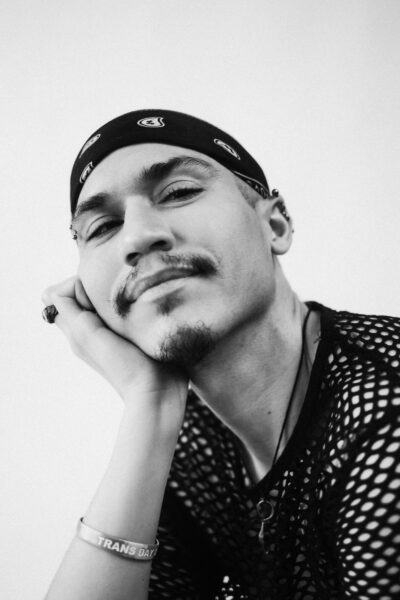 A person wearing a bandana, mesh shirt, and wristband rests their chin on their hand, looking at the camera with a slight smile.