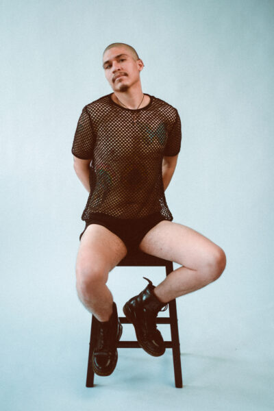 A person with short hair sits on a stool, wearing a black mesh shirt, black underwear, and black boots, against a plain light background.