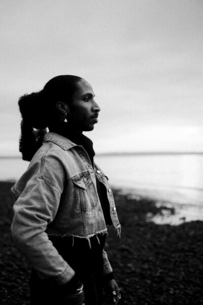 A person with long hair, wearing a denim jacket and earrings, stands on a rocky shore looking toward the water in a black and white photo.