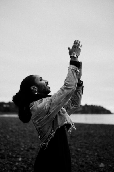 A person stands on a rocky shore, raising their hands upward with an expressive gesture, wearing a denim jacket and rings. The background shows water and a distant tree line.