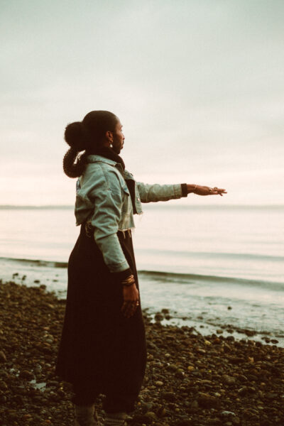 A person stands on a rocky shore facing the water, extending one arm outward, with overcast skies and calm waves in the background.