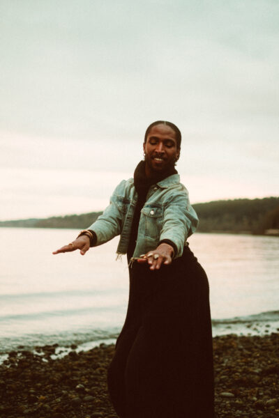 A person wearing a denim jacket and black skirt poses with outstretched arms on a rocky shoreline, with calm water and distant hills in the background.