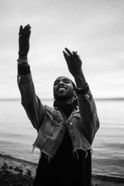 A person wearing a denim jacket stands near the water with arms raised, looking upward. The image is in black and white.