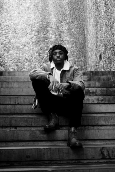 A person sits on outdoor steps with hands resting on knees, wearing boots, dark pants, and a jacket with a shearling collar; stone wall background.