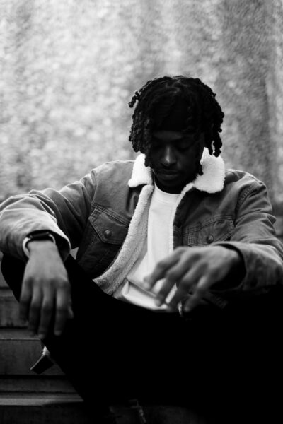A person with short dreadlocks, wearing a sherpa-lined jacket and white shirt, sits on steps while looking down; the image is in black and white.