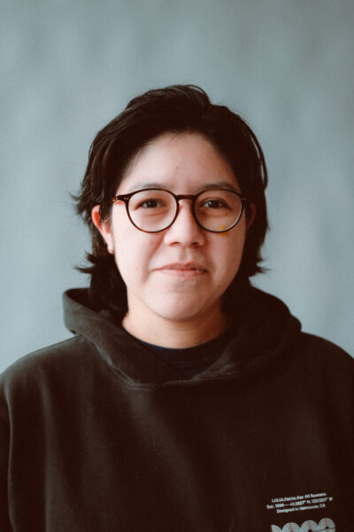 A person with short dark hair and glasses wearing a black hoodie stands in front of a plain light background, looking at the camera with a neutral expression.