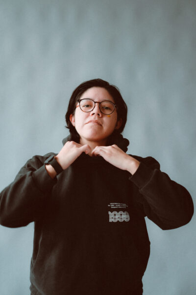 Person with short dark hair and glasses adjusts the collar of a black hoodie, standing against a plain, light gray background.