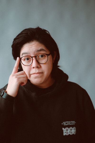 Person with medium-length black hair and glasses wearing a black sweatshirt points to their temple with a serious expression against a plain gray background.