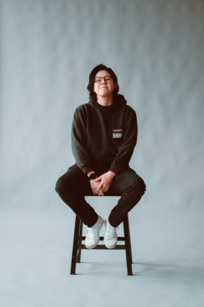 A person with glasses, wearing a dark hoodie, black pants, and white sneakers sits on a stool against a plain, light gray background.
