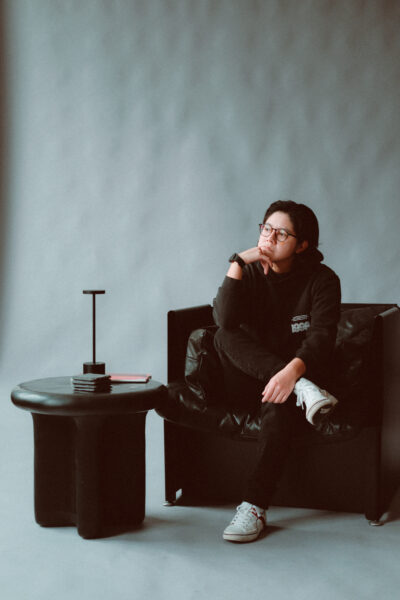 Person in glasses and dark clothing sits on a black chair with one leg crossed, resting their chin on their hand, beside a black table with books and a small lamp, against a plain background.