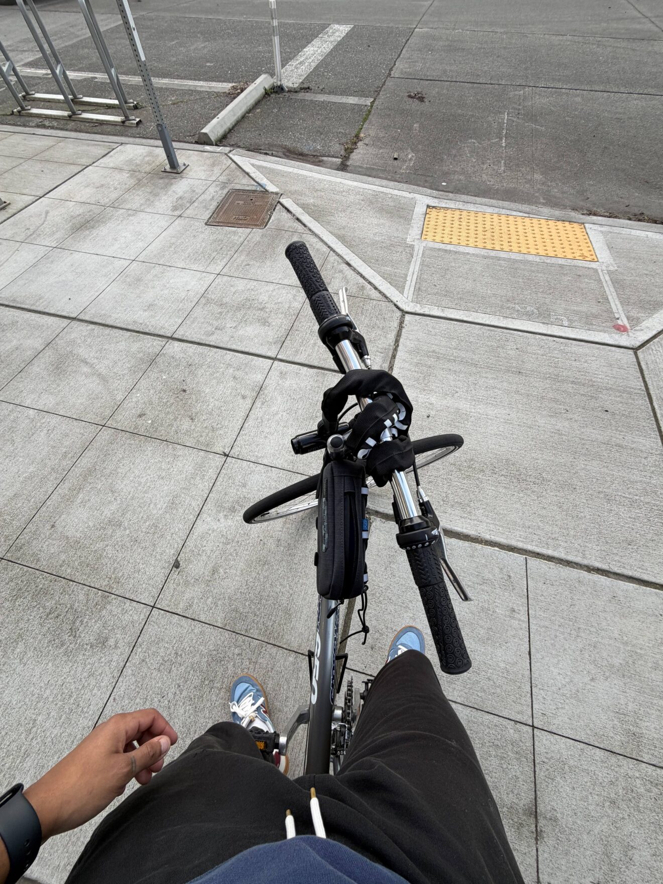 Person standing with a bicycle on a city sidewalk, showing bike handlebars, part of their legs, and the curb with a yellow tactile paving.