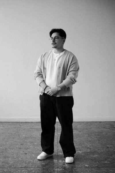 A man stands indoors against a plain wall, wearing glasses, a light cardigan, a white shirt, dark pants, and white shoes.