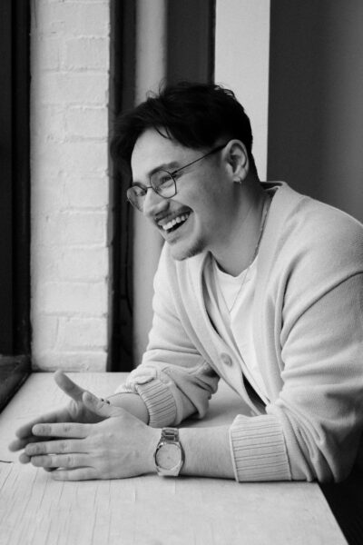 A man wearing glasses and a light cardigan sits by a window, smiling and leaning forward with his hands resting on a table.