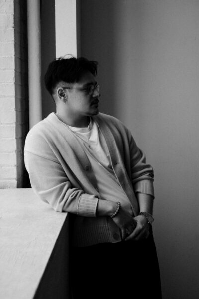 A person wearing glasses and a cardigan leans against a window ledge, looking to the side in a black and white setting.