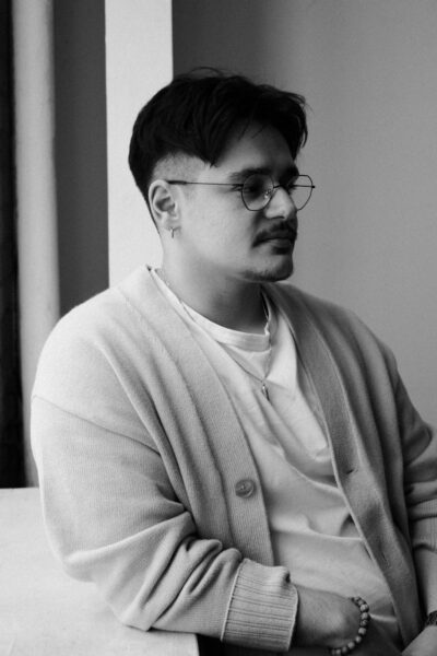 A man with glasses and facial hair wearing a light cardigan leans against a white surface, looking to the side in a softly lit room.