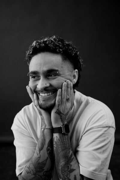 A person with curly hair, facial piercings, tattoos, and a smartwatch smiles while holding their face with both hands. The image is in black and white.