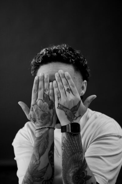 A person with curly hair and tattoos covers their face with their hands, wearing a watch and a bracelet, against a plain dark background.