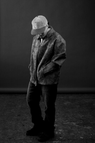 A person wearing a baseball cap, leather jacket, and jeans stands with hands in pockets, looking down, against a dark studio background.
