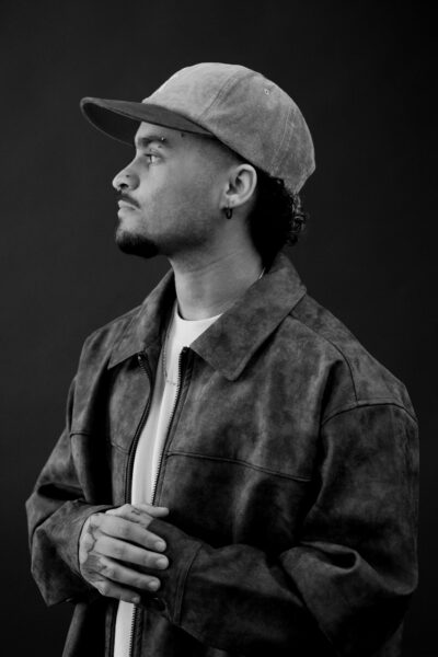 A man wearing a cap, earring, and suede jacket stands in profile against a dark background, looking to the left with hands clasped.