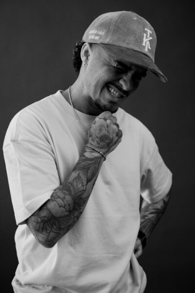 Man in a cap and t-shirt with tattooed arms stands against a dark background, clenching his fist near his face and wincing.