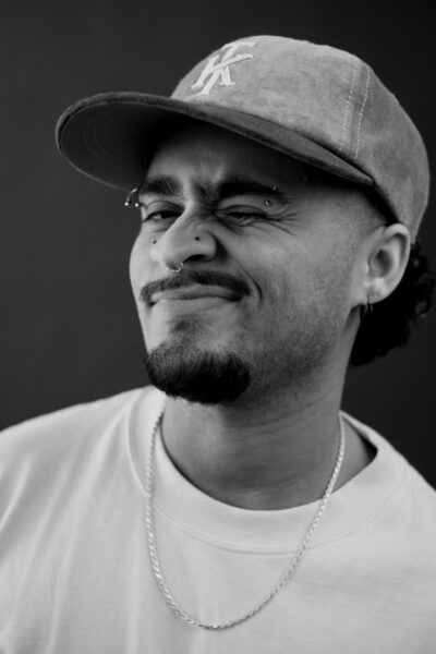 A person wearing a cap, chain necklace, and t-shirt is winking and smiling slightly. The image is in black and white.