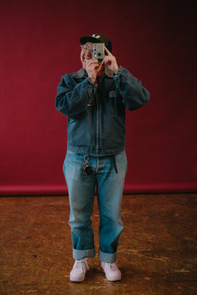 A person wearing a denim jacket, jeans, and pink sneakers stands on a brown floor against a red backdrop, holding a camera up to their face.