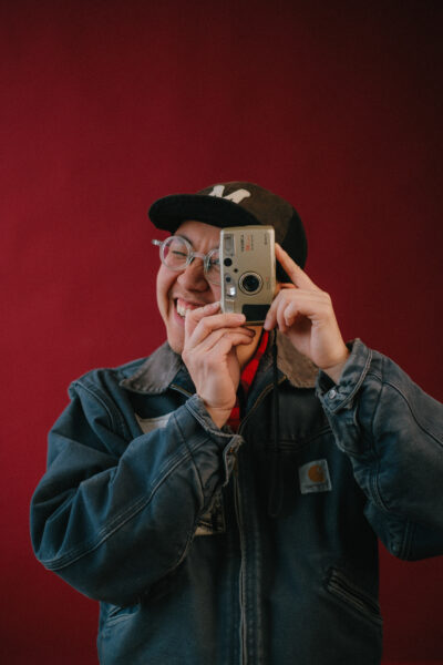 A person wearing glasses and a denim jacket smiles while holding a compact camera up to their eye against a plain red background.